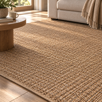 A natural woven jute area rug in a warm tan-brown color, styled in a bright, modern living room next to a wooden coffee table and a light beige sofa.