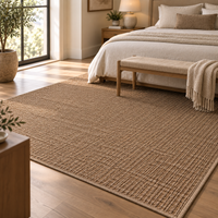 A large woven area rug in a warm tan-brown color, styled in a bright, modern bedroom with a wooden bed, bench, and light wood flooring.
