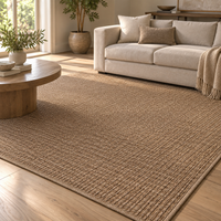 A large woven area rug in a warm natural tan color, shown in a bright, modern living room setting with a light beige sofa, a round wooden coffee table, and a potted plant.
